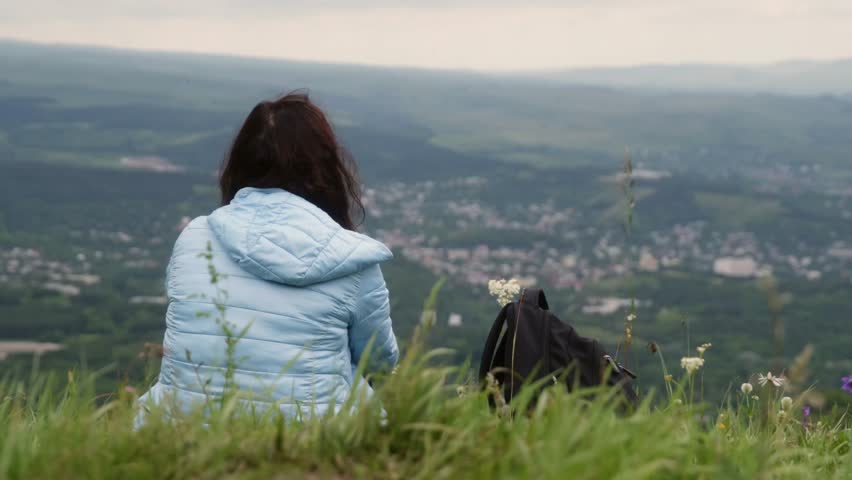 Woman and dog on hilltop overlooking valley, wearing light blue jacket, sitting in tall grass, calm reflective mood, distant town below, overcast sky, slow panoramic view