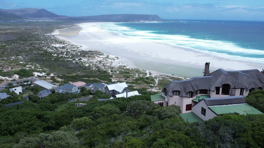 Aerial push-in shot gliding over luxury coastal residences towards the turquoise waves and white sands of Noordhoek Beach, South Africa.