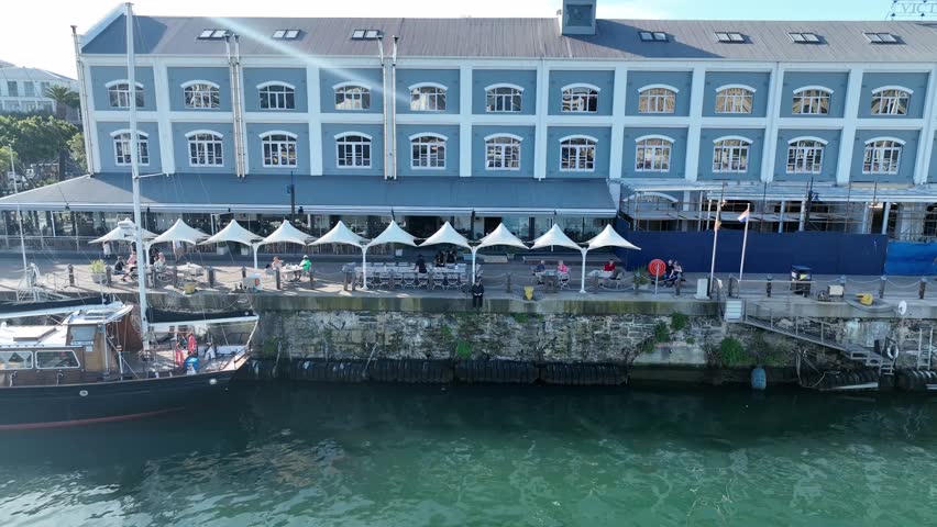 Aerial pull-out shot revealing the iconic V and A Waterfront in Cape Town, South Africa, featuring colonial architecture and a busy harbor under bright midday sun.