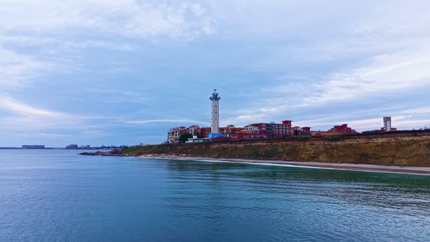 A coastal scene shows a lighthouse standing tall beside several buildings on cliffs by the water. The sky is cloudy and the sea is calm. This scene is captured in the afternoon.
