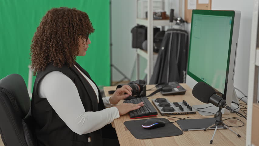 Woman at computer with hands on editing console and camera beside a green screen in a studio; concentration.