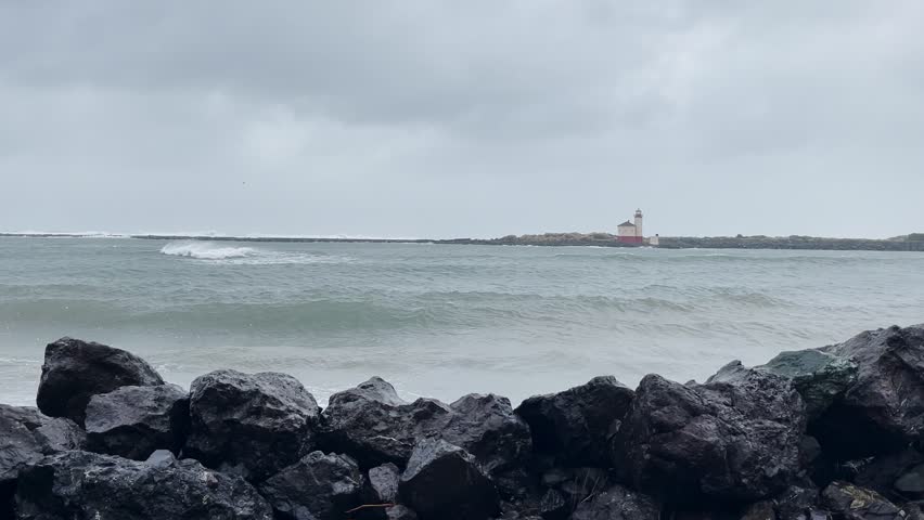 Ocean waves crashing against black rocks with distant lighthouse under grey sky on stormy day