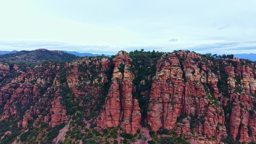 Rock formations in Sedona showcase red and brown colors under a cloudy sky. The landscape features trees and hills in the background, attracting hikers and nature lovers.