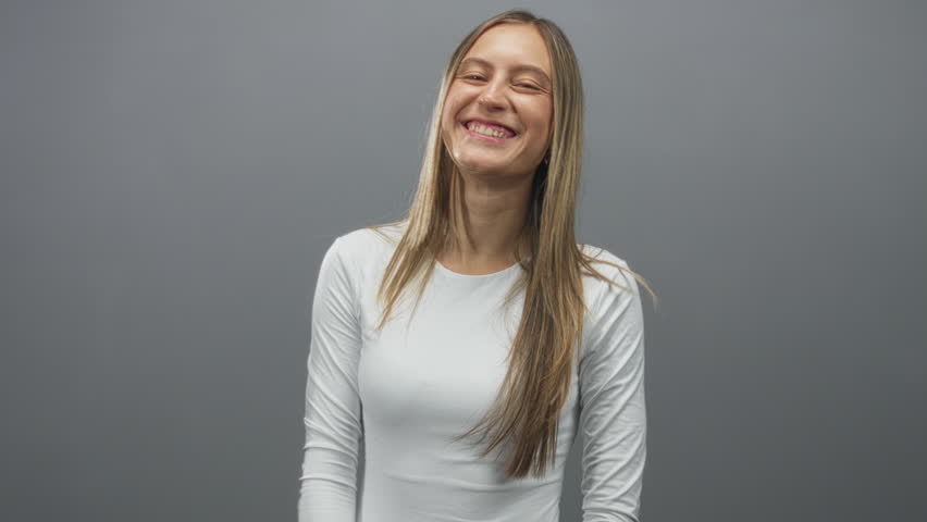 Young hispanic woman flashing rock horns hand gesture in gray studio wearing a white long sleeve top; joyful celebration.
