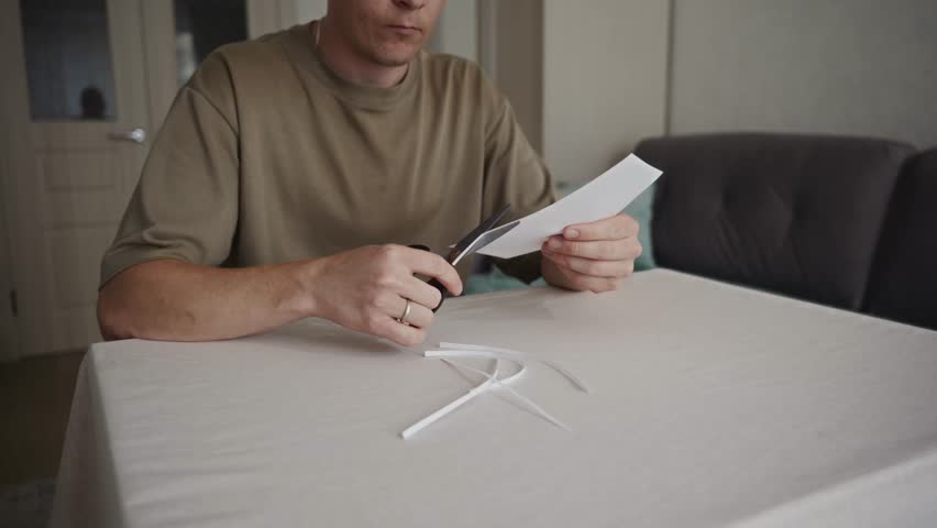 Man sitting at a table cutting white paper with scissors while small paper pieces lie on the table. Everyday indoor activity showing simple manual work. High quality 4k footage