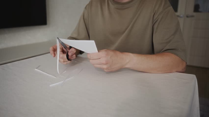 Man continues cutting a piece of white paper with scissors while sitting at a table. Paper strips accumulate on the table as the blades repeatedly close and open during the cutting process. 4k footage