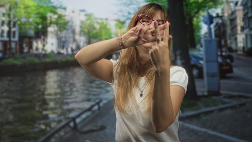 Woman with red glasses forming a hashtag with fingers while smiling on an amsterdam street canal walkway, hands forward and rings visible; playful connection.