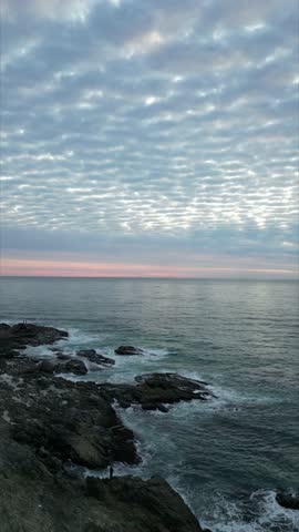 Vertical drone pullback over rocky coastline and ocean at sunrise with textured cloud pattern and gentle waves at Nambucca Heads, New South Wales, Australia. Calm coastal landscape