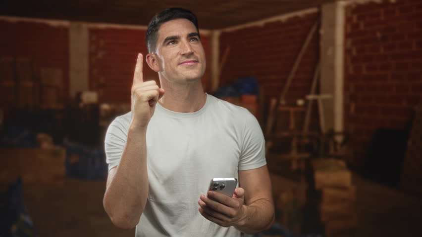 Man points finger upward while holding smartphone inside a building with brick walls and crates and ladders; optimism.