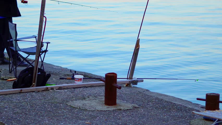 Male fisherman in black jacket casts line into water from pier with fishing gear visible
