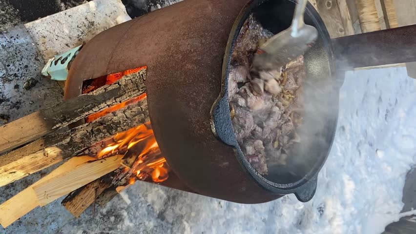 Large pot filled with meat cooking over an open fire, emitting steam and surrounded by snow, highlighting the outdoor cooking process