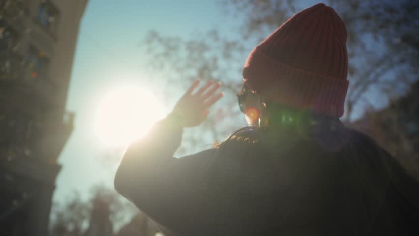 Back view of a woman standing in a busy city environment, raising her hand toward the sun. Footage captures the feeling of freedom, hope, and aspiration amidst the urban hustle. High quality 4k