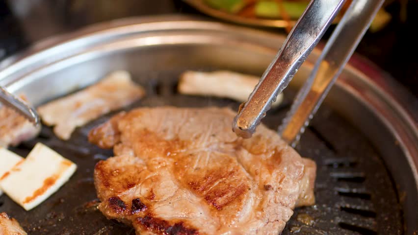 Metal tongs lift and flip a thick, seared pork steak on a circular tabletop grill.