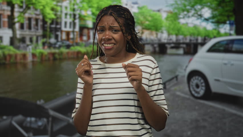 African american woman grimaces, fingers pinching her collar of a striped shirt on a canal street with parked car; disgust.