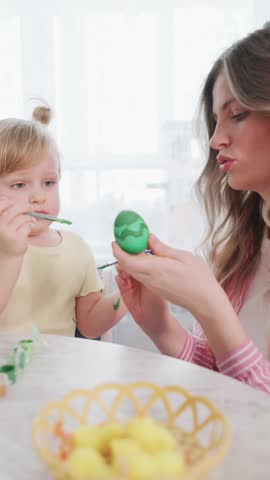 Mother and child paint Easter eggs together. Mother smiles, focused on decorating egg. Child concentrates, holding brush carefully. Bright table set with colorful eggs and baskets