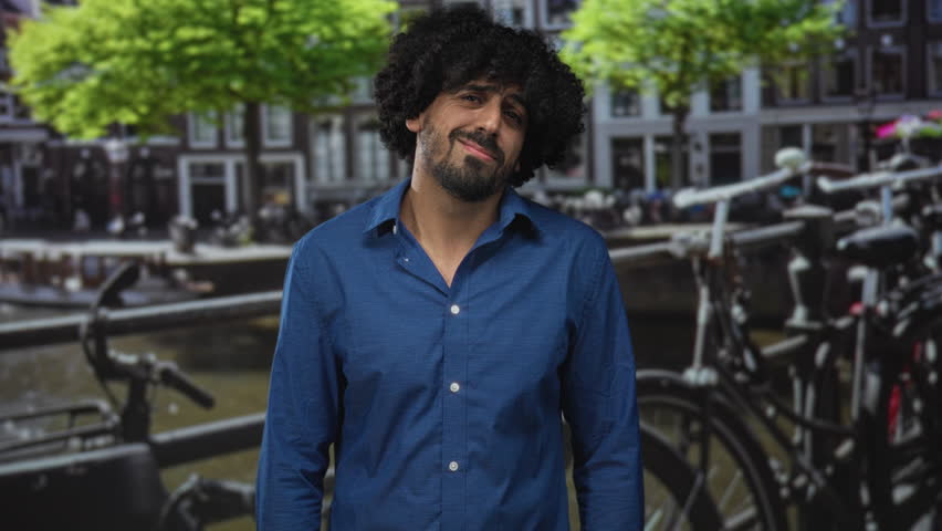 Man with curly hair wearing a blue shirt gesturing with hands on a canal street in amsterdam near bicycles; thoughtful reflection.