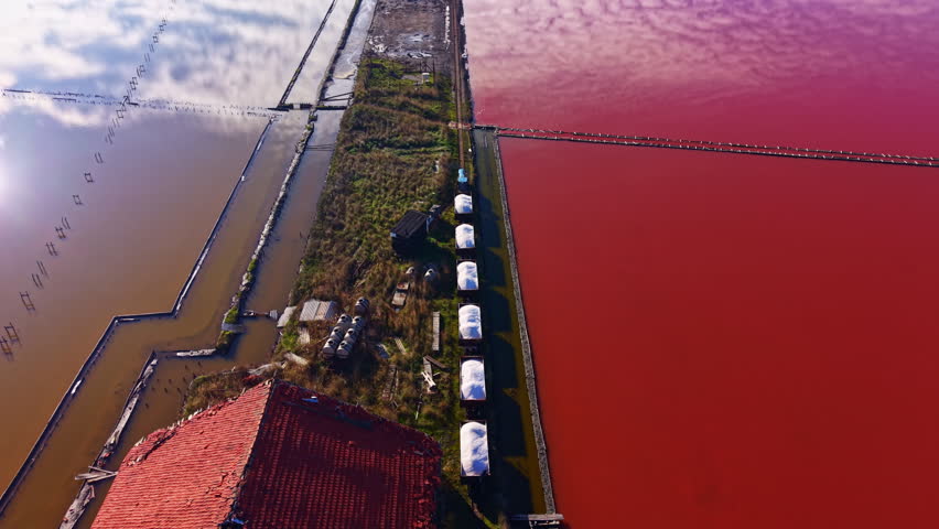 Aerial view of striking salt flats with vivid red and brown waters divided by a narrow, grassy path. Structures and equipment lie beside the flats under a blue sky, reflecting a unique landscape.