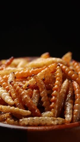 Fried bamboo worms or bamboo caterpillar in brown wooden bowl rotating slowly on balck background. Thai street food