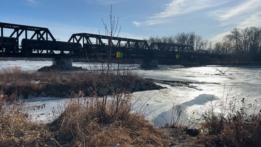 Cargo train tanks crossing bridge frozen river sunny day, Calgary, Alberta, Canada
