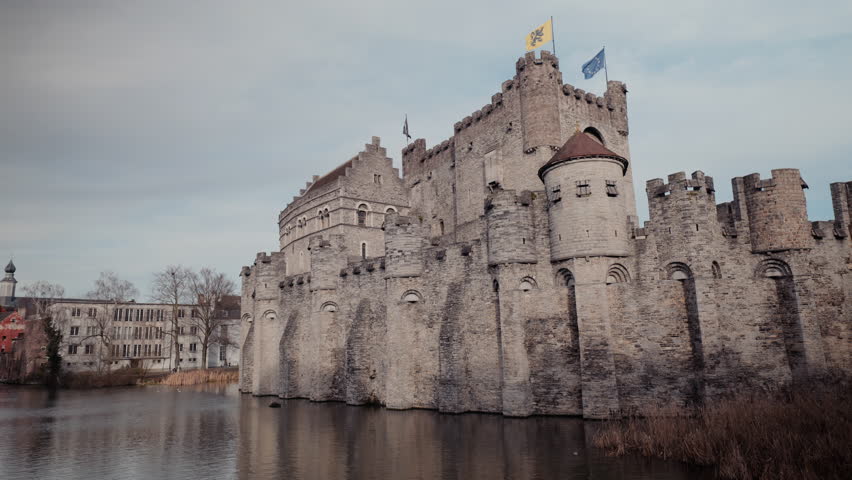 The imposing stone walls and round towers of the medieval Gravensteen castle rise above the moat in Ghent, Belgium, with Flemish and European flags flying from the battlements under a dramatic overcas