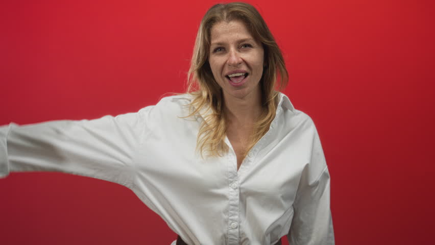 Woman in white shirt showing l sign with hand to forehead and open mouth in red studio; playful teasing defiance.