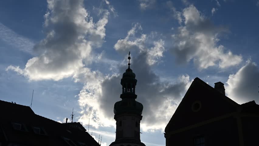 Time lapse of a historic church tower rising between old city rooftops under a dramatic sky filled with textured clouds.