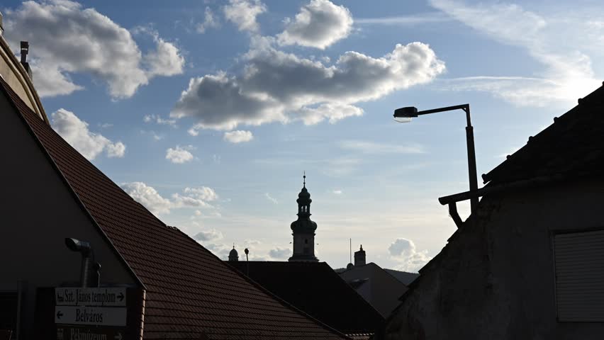 Time lapse View of a historic church tower rising above traditional European rooftops with a dramatic sky filled with scattered clouds.