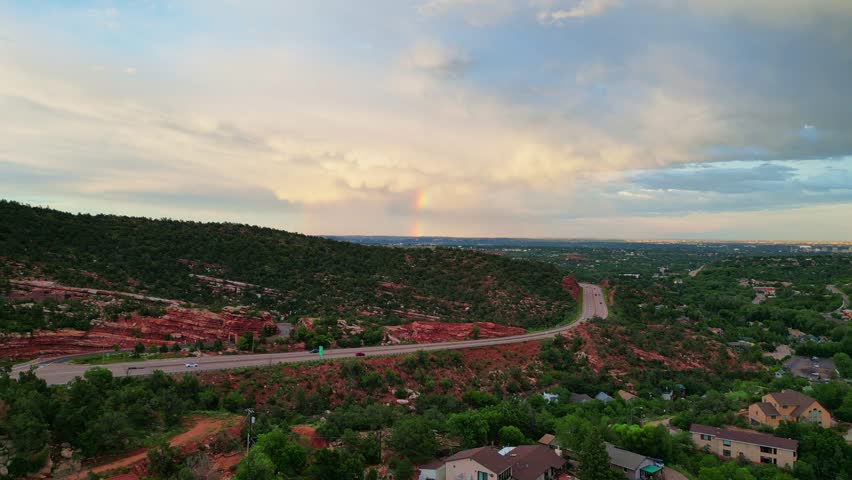 Wide aerial shot of a rainbow forming over a highway winding through red rock formations and green hills under a cloudy sunset sky. Estes Park, Colorado.