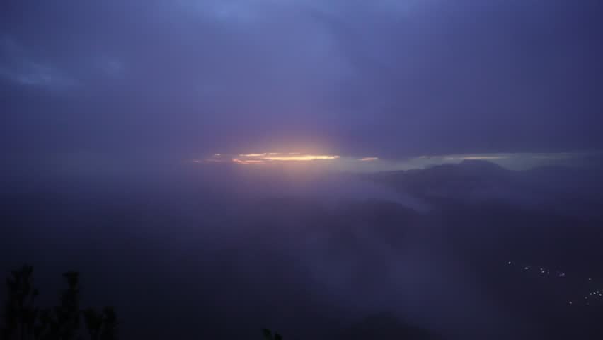 Moody sunset breaking through thick clouds above misty mountain silhouettes, with faint valley lights below.