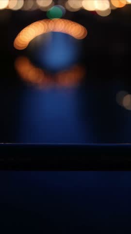 Vertical video of a classic Amsterdam canal at blue hour, starting with beautiful bokeh lights and pulling focus to reveal an illuminated historic arch bridge reflecting in the dark water, Netherlands.