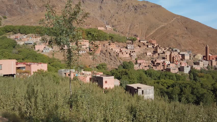 View of a traditional Berber hillside village near Imlil in Morocco’s High Atlas Mountains, likely Mzikene, surrounded by green orchards and rugged mountain slopes near Mount Toubkal.