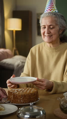 Vertical view of happy elderly lady wearing colourful conic hat holding white plate waiting for husband to serve piece of cake for her, then smelling it with excitement