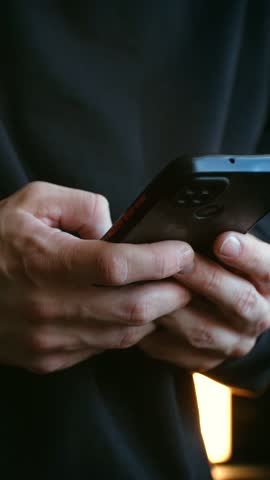 Close up male hands holding black smartphone, typing on touch screen, scrolling, swiping through social media feeds, applications, and messages in dimly lit room. Man hands typing on smartphone.