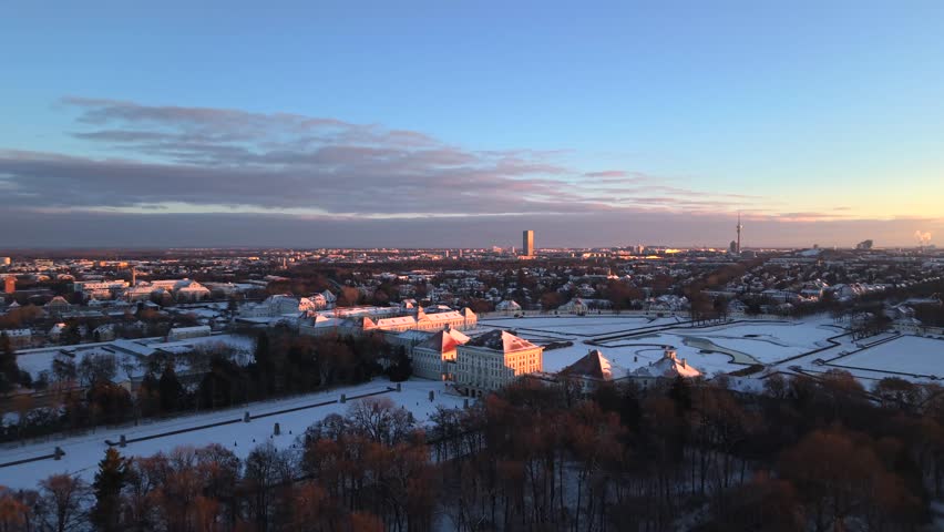 Aerial drone video of Nymphenburg Palace Park in Neuhausen-Nymphenburg, Munich, Bavaria, Germany. Fresh snow covers baroque palace gardens, canals and park at golden winter sunrise