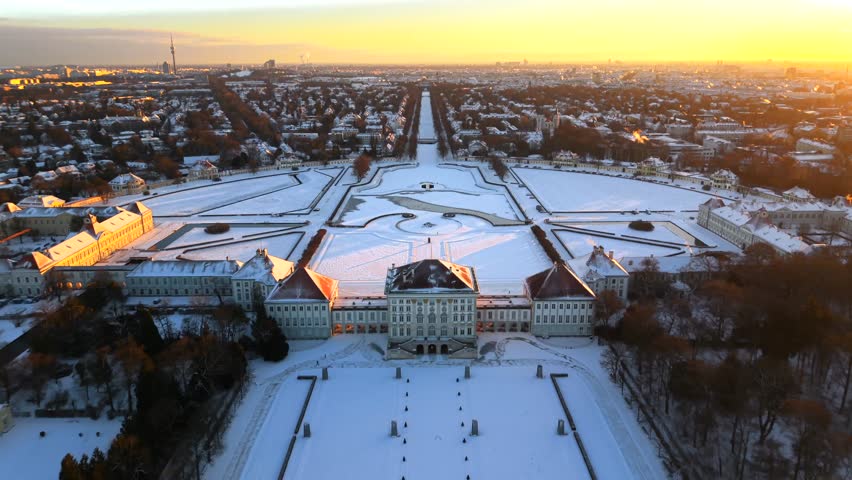 Cinematic aerial view of Nymphenburg Palace in Munich, Germany. Drone footage shows snow covered palace park, frozen canals and historic baroque architecture at golden dawn in January
