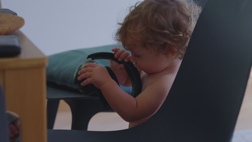 Toddler standing by chair holding black headphone earcup, inspecting it closely with focused expression during quiet and playful moment at home