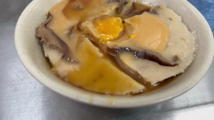 Taiwanese street food bowl kueh topped with shiitake mushrooms, runny egg yolk, and savory brown sauce served in a white bowl on metallic surface.

