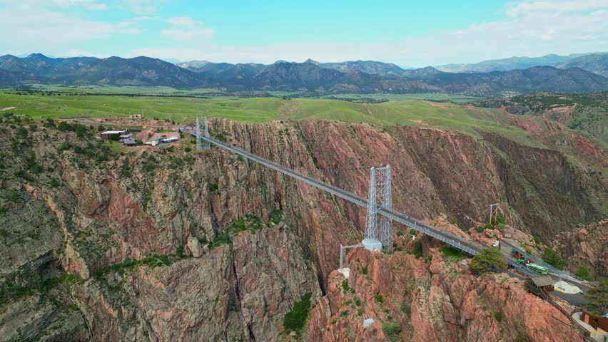 Wide aerial shot of the famous Royal Gorge Bridge spanning across a deep canyon in Colorado, surrounded by rugged mountains and rocky terrain under a blue sky.