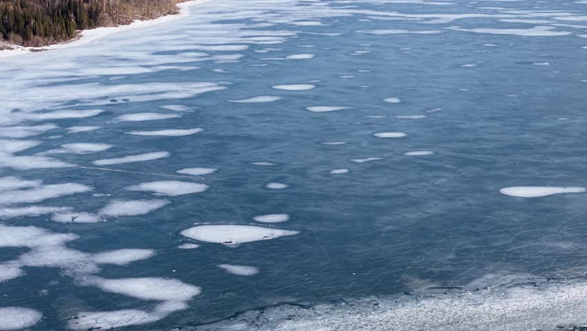 Aerial top-down view of patchy ice on a dark river or lake with irregular white floes forming circular shapes; filmed near Whitefish, Montana’s Flathead River.