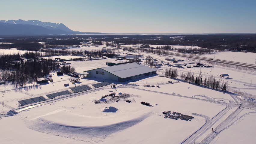 Aerial drone view of Alaska State Fairgrounds main exhibition hall in winter, snow-covered grounds and Chugach Mountains visible in background. 4K 60fps.