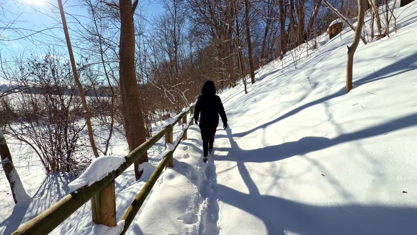 Person walking along snow-covered hillside path with wooden railing on Mežotne Hillfort Nature Trail, Latvia