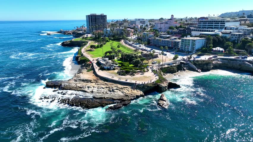 Aerial drone footage capturing the stunning La Jolla Cove in San Diego, California, featuring rugged cliffs, a lush green park with palm trees and crashing Pacific Ocean waves.