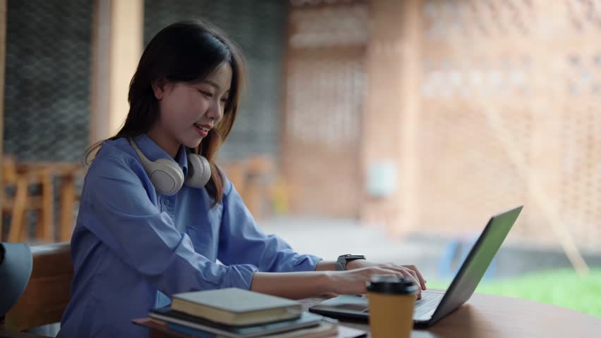 Asian woman focused on laptop computer for remote work or study. Young adult smiling in comfortable cafe setting. Digital task complete.