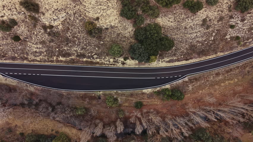 A winding road cuts through a landscape of dry earth and sparse trees. The view shows the road from above, highlighting the natural setting and the contrast of the ground and vegetation.