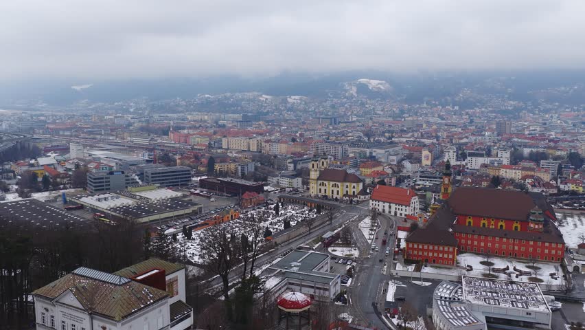 Aerial winter view of Innsbruck, Austria, shows a snow dusted city, a red roofed abbey, a pale yellow baroque church with twin towers, a railyard, trains, and roads.