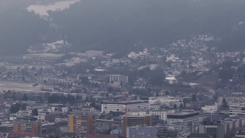 Aerial view of Innsbruck, Austria shows Nordkette slopes and Kranebitten airport as an airliner makes final approach, with winter light, haze, housing and cranes visible.
