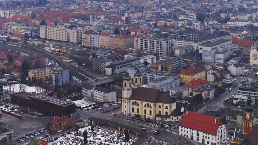 Aerial winter view in Innsbruck, Austria shows a Baroque twin tower church, a snow dusted cemetery, an arcaded walkway, rail lines, and trams and cars in motion.