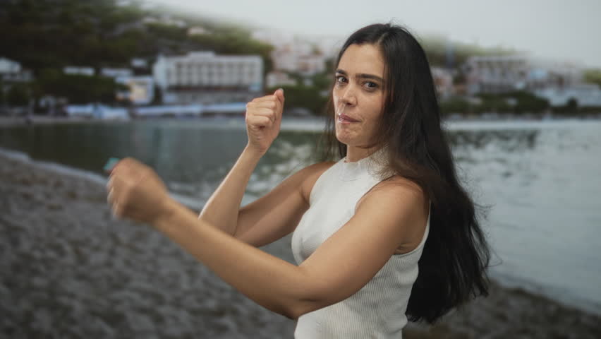 Woman with fists raised in a punching motion standing on a rocky seaside shore by a white building complex; determination.