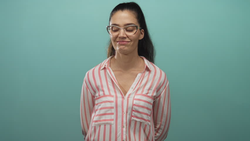 Woman rolls eyes in studio setting wearing glasses and a pink striped shirt with hands behind her back; skepticism.