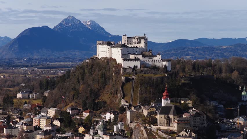 An aerial view over Salzburg, Austria, shows Hohensalzburg Fortress, Salzburg Cathedral domes, a red topped steeple, Untersberg, and Alpine ridges.